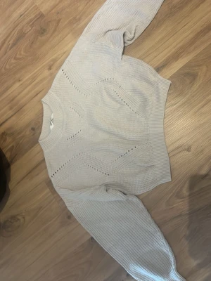 Ljusgrå cropped stickad tröja - Säljer en ljusgrå cropped stickad tröja med rund hals och dekorativa hålmönster framtill. Tröjan har långa ärmar och ribbade muddar vid ärmslut och nederkant. Perfekt för lager på lager och en chill look.Den sticks inte