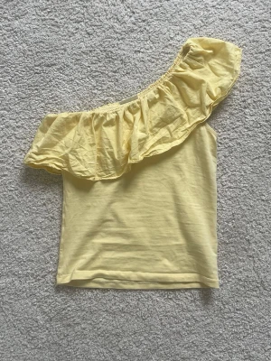 Gul one shoulder topp - Snygg ljusgul oneshoulder topp från Kappahl med volangdetalj längs halsringningen. Perfekt till en trendig, färgglad sommar look. Passar bra med jeans eller shorts. Storlek 146/152 men passar xs/xss Skriv vid intresse!!💞