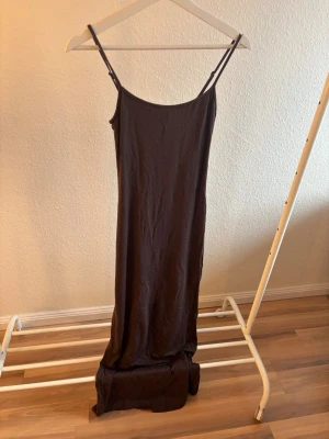 Brun maxiklännning med smala axelband - En stilren svart långklänning med smala justerbara axelband och rak passform. Klänningen har en enkel design utan mönster eller detaljer och är tillverkad i ett mjukt, tunt material som faller snyggt längs kroppen. Framhäver ens kurvor och passar perfekt till en stickad tröja eller skjorta på kyligare dagar eller bara med accessoarer på varmare dagar. Endast använd två gånger. Nypris 300 kr mitt pris 199 kr. 