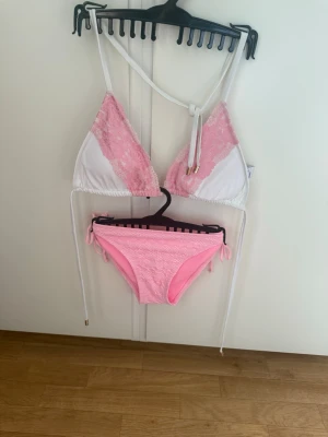 Vit och rosa bikini med spetsdetaljer - Säljer en söt bikini i vitt och ljusrosa med spetsdetaljer på överdelen och knyt i sidorna på underdelen. Bikinin har trekantsformad topp och matchande trosor, perfekt för sommaren och strandhäng. Överdel märket Elsa & Rosé från Bubbleroom o nederdel från Cubus helt nytt  utan pristaggar 