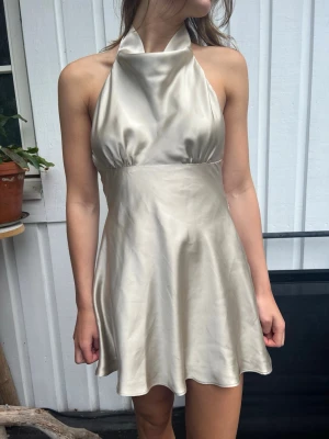 Champagnefärgad halterneckklänning - Snygg kort klänning i champagnefärgad satin med halterneck och öppen rygg. Klänningen har en stor rosett bak och ett elegant, slätt fall. Perfekt för dig som vill sticka ut med en stilren look.