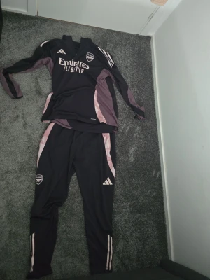Arsenal träningsset svart/rosa - Snyggt Arsenal träningsset från Adidas i svart med rosa och vita detaljer. Setet består av en långärmad tröja med klubbmärke och sponsorlogga samt matchande byxor med smal passform och klubbmärke på benet. Perfekt för fotbollsträning eller chill.