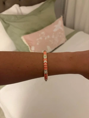 Färgglatt pärlarmband med blommor - Säljer ett färgglatt armband med små pärlor i orange, vitt och peach. Armbandet har blommönster och en rund form som ger en lekfull och trendig vibe. Perfekt accessoar för dig som vill addera lite färg till din outfit!