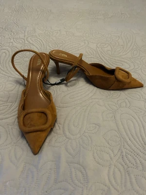 Bruna mocka-pumps med rem från ZARA - Snygga bruna pumps i mocka från ZARA med spetsig tå och dekorativ detalj framtill. Skorna har en smal rem runt hälen och låg klack, vilket ger en elegant och trendig look. 