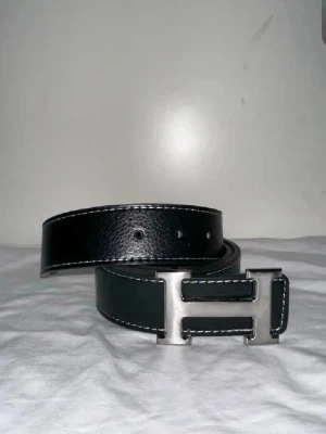 Svart bälte med silverfärgat H-spänne - Stilrent svart bälte med markerade vita sömmar och ett stort, silverfärgat H-format spänne. Bältet har en klassisk design och är tillverkat i skinn, vilket ger en lyxig känsla och passar till många olika outfits.