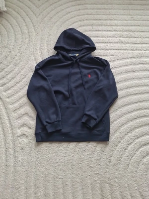 Mörkblå hoodie med röd logga - Snygg mörkblå hoodie från Polo Ralph Lauren med klassisk röd broderad logga på bröstet. Hoodien har känguruficka, dragsko i huvan och ribbade muddar vid ärmar och nederkant. Perfekt för en avslappnad och stilren look.