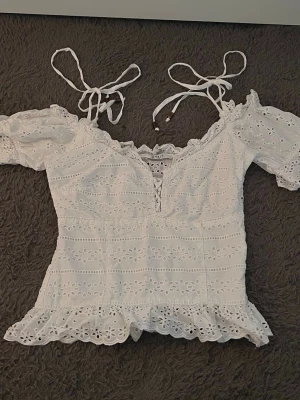Vit broderad topp med puffärm - Söt vit topp med broderade blommor, puffärmar och knytband vid axlarna. Djup v-ringning med snörning framtill och volangkant nedtill. Tillverkad i ett lätt bomullsliknande material som känns luftigt och somrigt. xs/s
