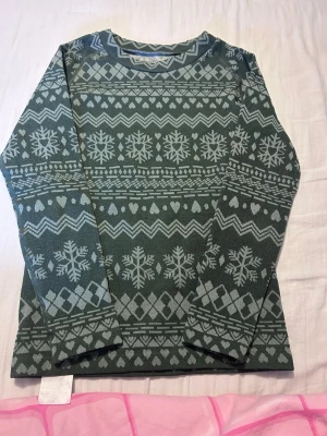 Grön stickad tröja med snöflingor - Mysig grön stickad tröja med ljusgrått vintermönster av snöflingor, hjärtan och zigzag-ränder. Tröjan har rund halsringning och långa ärmar. Perfekt för kalla dagar och ger en klassisk vinterlook.