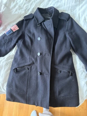 Svart ullrock med patch på ärmen - Stilren svart ullrock med dubbelknäppning och bred krage. Rocken har två snedställda fickor framtill och dekorativa axelklaffar. På vänster ärm sitter en patch med amerikansk flagga och siffran 6. Perfekt för dig som vill ha en klassisk men unik look.