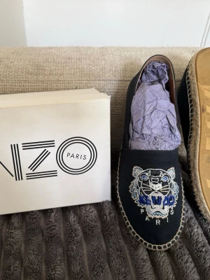 Svarta Kenzo loafers med tigerbrodyr - Säljer ett par mörkblå/svart espadrillos från Kenzo med ikonisk tigerbrodyr i vitt, blått och grått framtill. Skorna har naturfärgad flätad sula och är tillverkade i tyg med läderdetaljer på insidan. Säljer då dem är för små