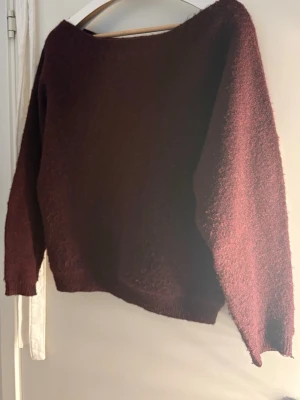 Mörkröd stickad tröja från Nelly - Mysig mörkröd stickad tröja från Nelly med lös passform och stor halsringning som gör att man kan ha den off shoulder. Tröjan har långa ärmar och är gjord i ett fluffigt, mjukt material som känns skönt mot huden. Perfekt för kyliga dagar och enkel att matcha till olika outfits.