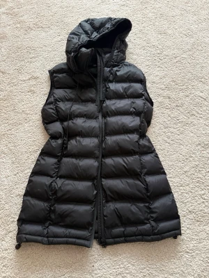 Svart lång dunväst med huva ZARA - Snygg svart lång dunväst med huva och dragkedja framtill. Västen har quiltad design, dragsko i huvan och är tillverkad i ett glansigt material. Perfekt för lager-på-lager och ger en cool streetwear-look.
