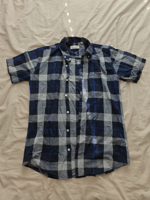 Burberry skjorta - Säljer en blårutig skjorta med korta ärmar och klassisk krage. Skjortan har vita knappar framtill och ett broderat märke på bröstet. Tillverkad i mjuk bomull, perfekt för en avslappnad stil.