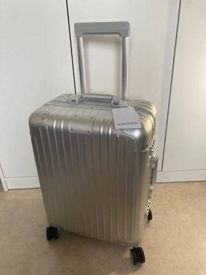 Silver resväska från Rimowa - Snygg silverfärgad resväska från Rimowa i aluminium med klassisk räfflad design. Väskan har fyra hjul, teleskophandtag och sidohandtag för enkel hantering. Perfekt för dig som vill resa med stil och funktion. Sista bilden är det en skada som hände under resan inget seriöst .