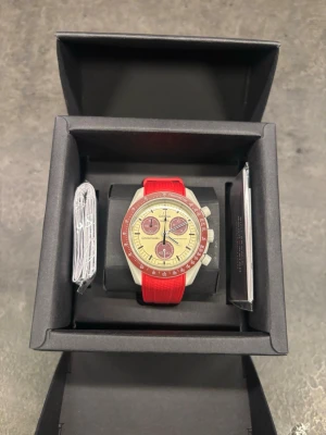 Omega Speedmaster med röd silikonrem - Snygg Omega Speedmaster klocka med röd silikonrem och beige urtavla. Klockan har röda detaljer runt urtavlan och på kronograferna. Klassisk design med sportig känsla och tydliga visare. Perfekt accessoar för dig som gillar stilrena och färgstarka klockor.