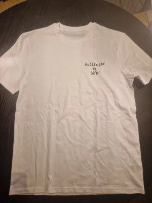 Vit t-shirt med Bullerbyn-tryck - Säljer en vit t-shirt med tryck fram och bak. På bröstet står det 'Bullerbyn är död!' och på ryggen finns ett stort färgglatt motiv med texten 'SIMON SUPERTI'. T-shirten har rund hals och korta ärmar. Perfekt för dig som gillar unika prints och streetwear.