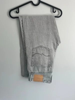 Grå raka jeans med bakficka - Säljer ett par ljusgrå jeans med rak passform och klassisk femficksdesign. Jeansen har en tydlig bakficka med dekorativ söm och en läderpatch vid linningen. Materialet är kraftig denim i bomull, perfekta för en avslappnad och trendig look.