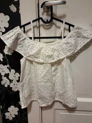 Vit off shoulder topp med broderi - Söt vit off shoulder topp med tunna axelband och volangkant. Toppen har broderade hålmönster över hela tyget och är gjord i ett lätt och luftigt material, perfekt för sommaren. Passar till jeans eller kjol för en fräsch look.