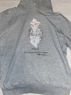 Grå hoodie Polo Bear Ralph Lauren - Säljer en grå hoodie från Ralph Lauren med den ikoniska Polo Bear-figuren tryckt på framsidan. Hoodien har huva och ribbade muddar. Perfekt för dig som gillar klassisk streetwear med en twist. Materialet är mjukt och skönt, troligen bomullsblandning.