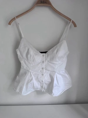 Vit korsettopp med knappar - Säljer en vit korsettopp med smala axelband och knappar framtill. Toppen har en figurnära passform upptill och en lätt utsvängd nederdel. Perfekt för dig som vill ha en trendig och stilren look i sommar.