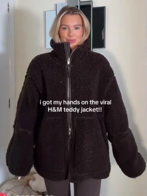 Mörkbrun virala H&M teddyjacka med dragkedja - Mysig svart teddyjacka från H&M med hög krage och grov dragkedja framtill. Jackan har två stora fickor och en oversized passform som ger en trendig look. Perfekt för kyliga dagar när du vill vara både varm och snygg.