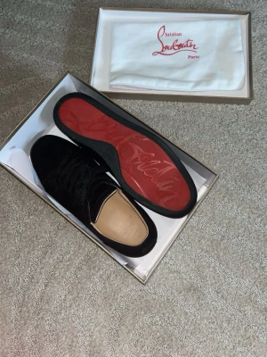Svarta sneakers från Christian Louboutin - Snygga svarta sneakers från Christian Louboutin i mocka med klassisk röd sula och svarta skosnören. Skorna har en stilren design och diskreta detaljer, perfekt för dig som vill ha något exklusivt och trendigt.