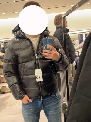 Svart pufferjacka med huva - Säljer en svart pufferjacka med glansig finish och stor huva. Jackan har dragkedja framtill, två sidofickor och breda quiltade paneler som ger en riktigt puffig look. Perfekt för kalla vinterdagar och riktigt snygg till jeans eller sweatpants.