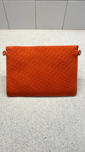 Orange kuvertväska med präglat mönster - Snygg orange kuvertväska i läderimitation med präglat monogrammönster över hela ytan. Väskan har avtagbara och justerbara axelremmar i matchande färg, vilket gör den flexibel att bära både som clutch och crossbody. Perfekt för dig som vill sticka ut med färg.