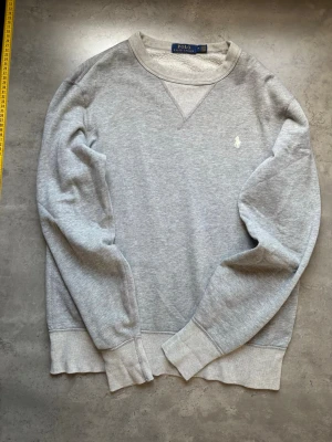 Grå sweatshirt från Polo Ralph Lauren - Klassisk grå sweatshirt från Polo Ralph Lauren med rund halsringning och vit broderad logga på bröstet. Tröjan har ribbade muddar vid ärmslut och nederkant samt en snygg detalj vid halsen. Perfekt för en avslappnad och stilren look.