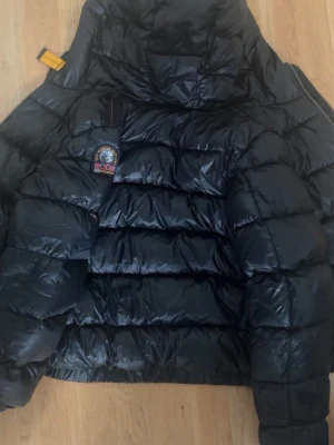 Parajumpers jacka  - Snygg och varm vinterjacka från Parajumpers med klassisk puffer-design. Jackan har en stor och mysig huva, tjock dunvaddering och den ikoniska Parajumpers-loggan på ärmen. Den är svart med grått innerfoder och har flera detaljer i den klassiska Parajumpers-stilen.  Jackan är i Y-M storlek och passar perfekt för kalla höst- och vinterdagar. Klassisk modell som är både stilren och varm. Äkta!