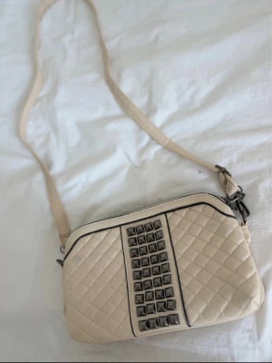 Beige axelväska med nitar - Snygg beige axelväska i quiltat konstläder med silverfärgade nitar framtill. Väskan har justerbar axelrem och dragkedja upptill. Perfekt för dig som gillar en edgy men stilren look.