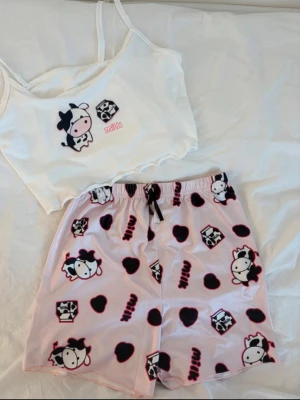 Söt pyjamas med ko- och mjölktryck - Tvådelad pyjamas med vitt linne och ljusrosa shorts. Linnet har smala axelband och tryck av en gullig ko, mjölkpaket och texten 'milk'. Shortsen har elastisk midja och matchande tryck med kor, hjärtan och mjölkpaket. Perfekt för mysiga hemmakvällar. Storleken på tröjan och mjukisarna är inte detsamma då byxorna är betydligt mycke mindre en toppen 