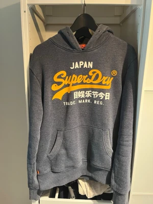 Blå Superdry hoodie med tryck - Säljer en blå hoodie från Superdry med stort gult och vitt tryck på bröstet och nr 92 på armen. Klassisk känguruficka framtill och huva med dragsko. Mjuk insida och ribbade muddar vid ärmar och nederkant. Perfekt för en avslappnad streetwear-look.