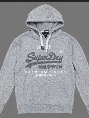 Grå hoodie från Superdry - Snygg gråmelerad hoodie från Superdry med stor logga och japansk text på bröstet. Klassisk känguruficka framtill, dragsko i huvan och mjukt material som känns skönt mot huden. Perfekt för en avslappnad streetstyle-look.