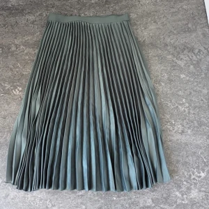 Lång plisserad kjol i grå satin - Midikjol i blå satin med plisserad design. Kjolen har ett brett midjeband.