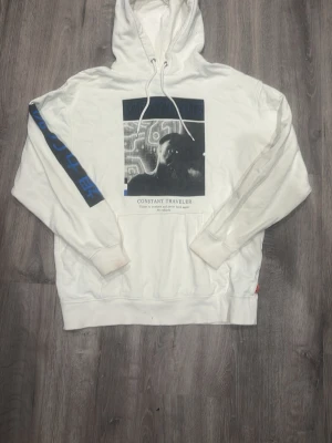 Valient  - Snygg vit hoodie med huva och dragsnören. Framsidan har ett svartvitt grafiskt tryck och texten 'CONSTANT TRAVELER'. Ena ärmen har blå text och en diagonal blå linje. Tillverkad i mjukt bomullsmaterial, liten fläck på ena mudden på armen 