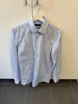 Ljusblå skjorta från Hugo Boss, stl 38 - Snygg ljusblå skjorta från Hugo Boss i slim fit-modell. Klassisk krage, knäppning framtill och långa ärmar. Perfekt för dig som gillar stilrena och enkla plagg. Skjortan är tillverkad i bomull för en skön känsla.