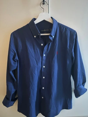 Marinblå skjorta från Ralph Lauren - Klassisk marinblå skjorta från Ralph Lauren med knappslå och vit knappdetalj. Skjortan har lång ärm, button-down krage och den ikoniska röda logotypen broderad på bröstet. Tillverkad i bomull för en skön och stilren look.