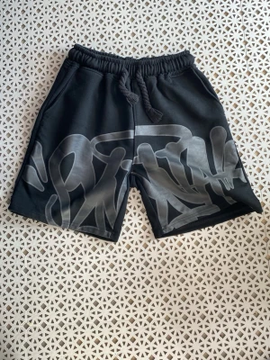 Svarta syna shorts  - Säljer ett par svarta shorts med grått graffiti-inspirerat tryck framtill. Shortsen har resårmidja med snörning och sidofickor samt en bakficka. Perfekta för en avslappnad streetwear-look.