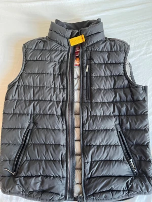 Grå dunväst med dragkedjor - Snygg svart dunväst från Parajumpers med quiltad design och hög krage. Västen har två framfickor med dragkedja och en gul detalj vid halsen. Perfekt för lager-på-lager och ger en sportig look.