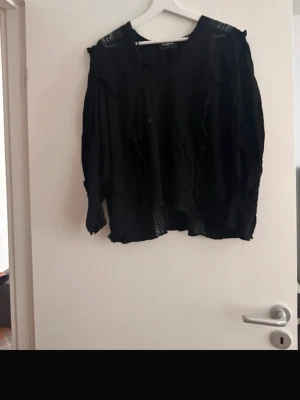 Svart blus från The Kooples - Snygg svart blus från The Kooples med avslappnad passform och tunna, genomskinliga detaljer. Blusen har långa ärmar och en lös, lite bohemisk look med dekorativa band vid halsen. Perfekt för dig som gillar en edgy och trendig stil.