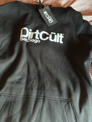 Svart Dirt Cult hoodie med tryck - Säljer en svart hoodie från Dirt Cult med stor vit logga och texten 'San Diego' på bröstet. Den är helt oanvänd, säljer den för den var fel storlek, den är i storlek L i herrstorlek, skriv vid mer frågor💓