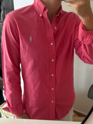 Rosa skjorta från Ralph Lauren - Säljer en snygg rosa skjorta från Ralph Lauren med klassisk button-down krage och vita knappar. Skjortan har det ikoniska broderade logotypen på bröstet och är tillverkad i bomull. Perfekt för dig som vill ha en stilren och färgglad look. Liten tråddragning vid axeln därav priset 