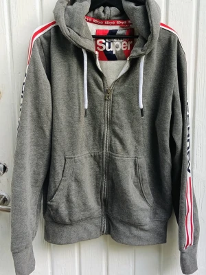 Superdry Hoodie  - Grå hoodie med dragkedja från Superdry. Huva med vita snören och stora fickor framtill. Ärmarna har vita och röda ränder samt Superdry-logga längs hela armen. Mjuk bomullsmix och ribbade muddar. Stl xxl, endast använd ett fåtal gånger