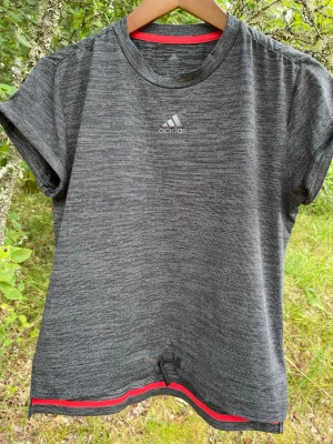 Grå tränings-tshirt från Adidas - Snygg gråmelerad tränings-tshirt från Adidas med korta ärmar och rund hals. T-shirten har diskret Adidas-logga på bröstet och röda detaljer vid nederkanten. Tillverkad i ett lätt och snabbtorkande funktionsmaterial som passar perfekt till träning.