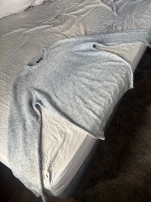 Ljusgrå stickad tröja - Mysig ljusgrå stickad tröja med rund hals och långa ärmar. Tröjan har en enkel och stilren design, perfekt för lager på lager och chill dagar. Materialet ser mjukt och fluffigt ut, vilket gör den extra skön att bära.