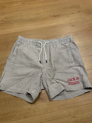 Randiga badbyxor från Jack & Jones - Säljer ett par randiga shorts från Jack & Jones i vitt och grått med snörning i midjan och fickor på sidorna. Shortsen har en broderad logga i rött på vänster ben och är tillverkade i ett lätt bomullsmaterial. Perfekta för varma dagar!