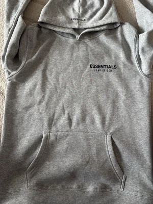 Grå hoodie Essentials Fear of God - Snygg grå hoodie från Essentials Fear of God med svart logga på bröstet. Klassisk känguruficka framtill och huva. Tillverkad i mjukt bomullsmaterial som känns skönt mot huden. Perfekt för en avslappnad och trendig streetwear-look.