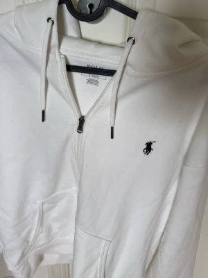 Vit Ralph Lauren Zip Hoodie - Ralph Lauren Vit Zip Hoodie  Äkta Ralph Lauren zip hoodie i mycket fint skick. En stilren och klassisk hoodie med bekväm passform, perfekt för vardagsbruk. Den vita färgen gör den enkel att matcha med både jeans och andra plagg.  * Märke: Ralph Lauren * Färg: Vit * Modell: Zip hoodie * Skick: Mycket fint skick * Klassisk design * 100% äkta * Storlek: M Skicka gärna ett meddelande om du har frågor eller vill ha fler bilder.