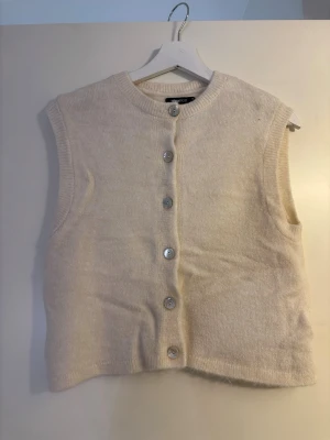 Beige stickad väst med knappar - Säljer en beige stickad väst med rund hals och snygga knappar framtill. Västen har en enkel och clean look som passar till många olika outfits. Perfekt för lager på lager och ger en soft vibe till din stil.